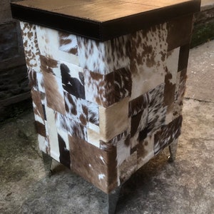 May include: A square ottoman with a brown leather top and a patchwork design of cowhide in shades of white, brown, and black. The ottoman has four silver legs.