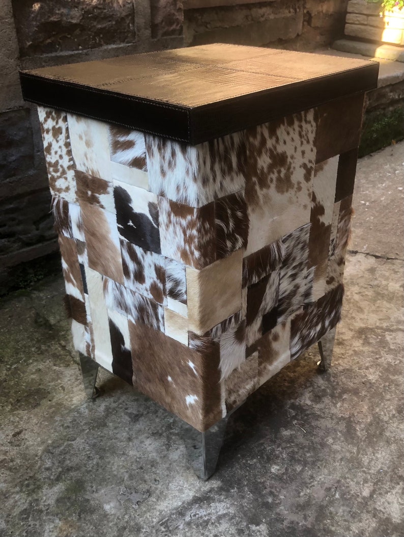 May include: A square ottoman with a brown leather top and a patchwork design of cowhide in shades of white, brown, and black. The ottoman has four silver legs.