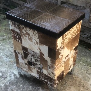 May include: A brown leather-topped storage ottoman with a patchwork design of cowhide in various shades of brown, white, and black. The ottoman has four silver metal legs.