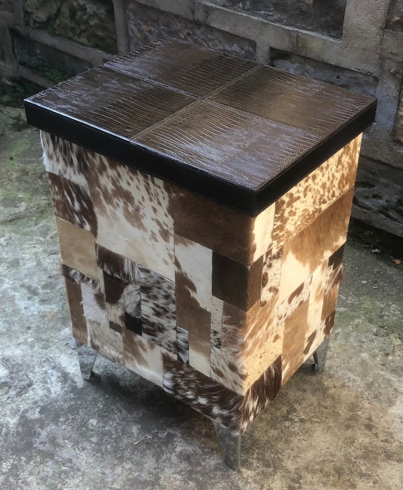May include: A brown leather-topped storage ottoman with a patchwork design of cowhide in various shades of brown, white, and black. The ottoman has four silver metal legs.