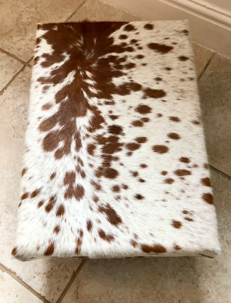 Puede incluir: Un otomano de piel de vaca blanca y marr&oacute;n con un patr&oacute;n de manchas marrones y blancas.
