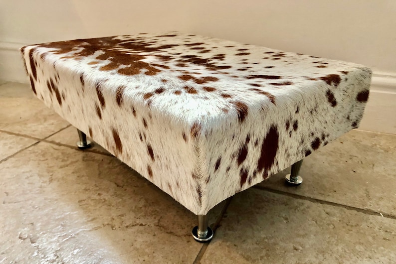 Puede incluir: Un reposapi&eacute;s de piel de vaca blanca y marr&oacute;n con cuatro patas de metal plateado.
