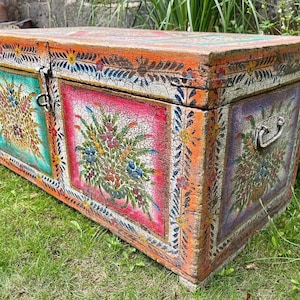 May include: An antique wooden chest with a distressed, painted finish. The chest features floral designs in pink, green, and purple, with orange and blue accents. It has a metal latch and handle. The chest is on a grassy surface.