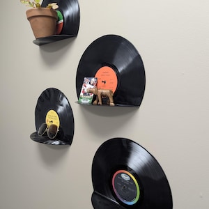 May include: Four black vinyl record shelves mounted on a wall, each holding a different item. One shelf holds a small potted plant, another has sunglasses, and a third displays a wooden figurine and a card. The fourth shelf is empty.