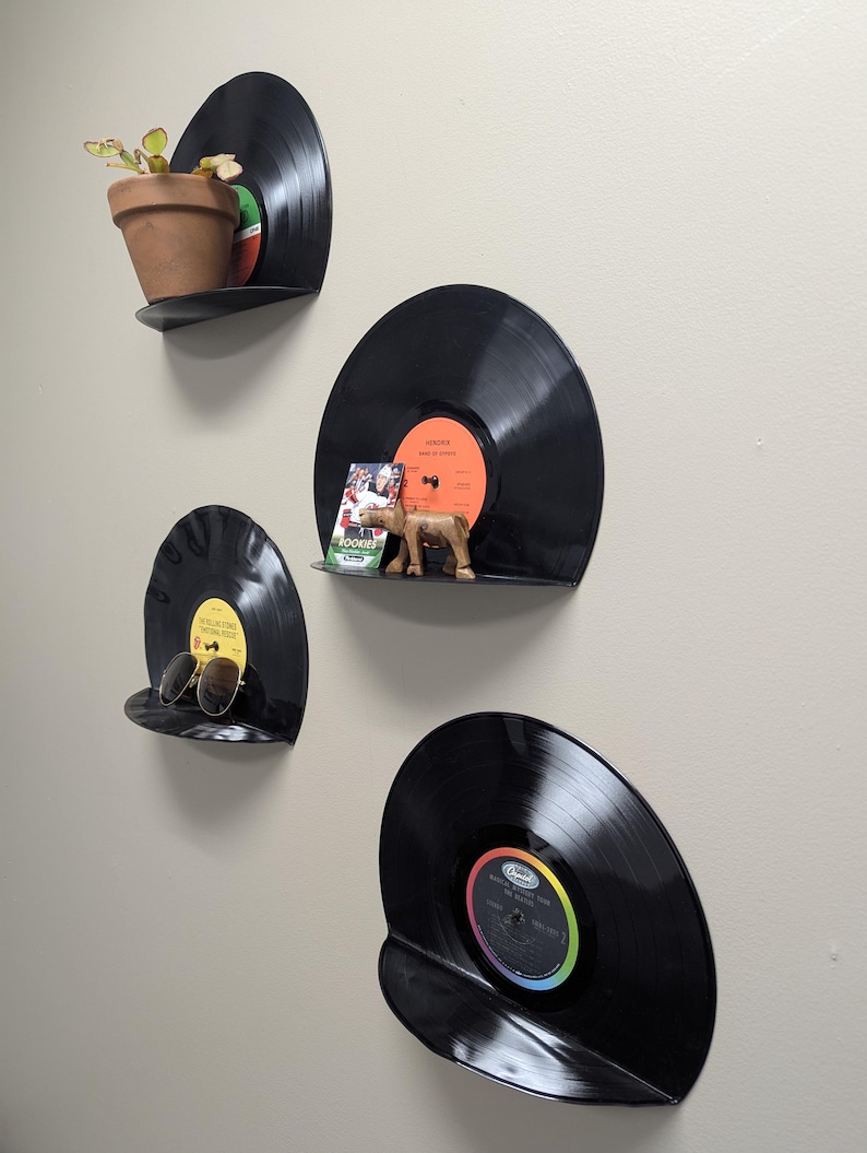 May include: Four black vinyl record shelves mounted on a wall, each holding a different item. One shelf holds a small potted plant, another has sunglasses, and a third displays a wooden figurine and a card. The fourth shelf is empty.