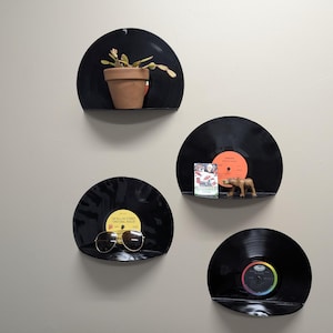May include: Four black vinyl record shelves mounted on a light gray wall. Each shelf holds a different item: a potted plant, a small figurine, sunglasses, and a record. The shelves are arranged in a vertical pattern.
