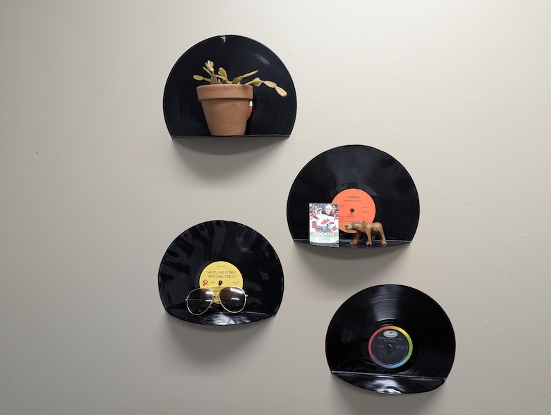 May include: Four black vinyl record shelves mounted on a light gray wall. Each shelf holds a different item: a potted plant, a small figurine, sunglasses, and a record. The shelves are arranged in a vertical pattern.