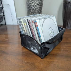 May include: A black, upcycled vinyl record CD holder filled with CDs. The holder is made from a repurposed record and sits on a wooden surface. The CDs are in clear plastic cases, with various album art visible.