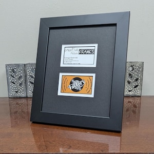 May include: A black picture frame with two business cards inside. The top card reads "creative Picture FRAMES" and the bottom card says "Factory Records". The frame is on a wooden surface, with decorative metal objects on either side.