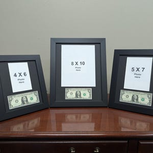May include: Three black picture frames with a dollar bill inside each frame. The frames are 4x6, 5x7, and 8x10 inches. The frames are on a wooden table.