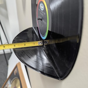 May include: A black vinyl record shelf, approximately 3 inches deep, mounted on a wall. The record is labeled "Magical Mystery Tour The Beatles". A measuring tape shows the shelf's depth.