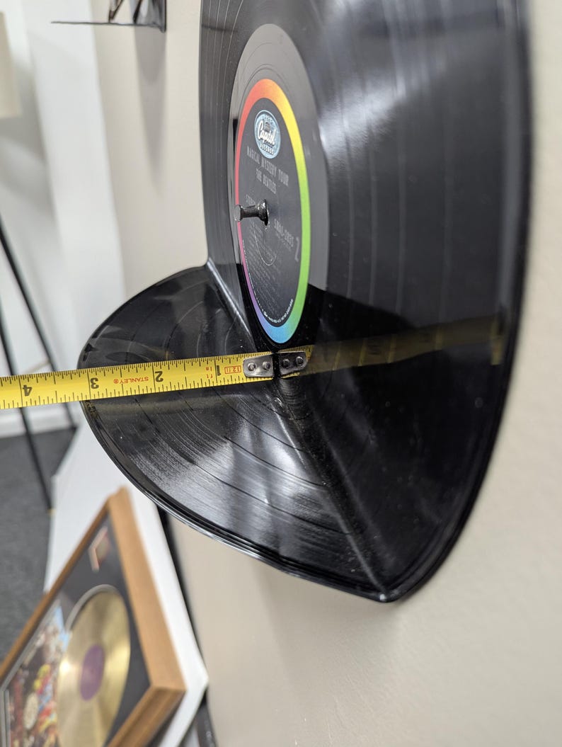 May include: A black vinyl record shelf, approximately 3 inches deep, mounted on a wall. The record is labeled "Magical Mystery Tour The Beatles". A measuring tape shows the shelf's depth.