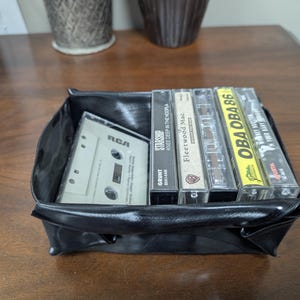 May include: A black, square, vinyl tray filled with vintage cassette tapes. The tapes include titles like "Starship," "Fleetwood Mac," and "OBA OBA 86." One tape is labeled "RCA."