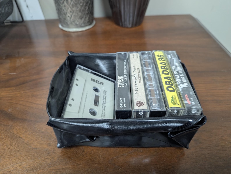May include: A black, square, vinyl tray filled with vintage cassette tapes. The tapes include titles like "Starship," "Fleetwood Mac," and "OBA OBA 86." One tape is labeled "RCA."