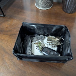 May include: A black, glossy, rectangular catch-all tray holding keys, coins, a folding knife, and a small decorative plate. The tray sits on a wooden surface, providing a convenient storage solution for everyday items.