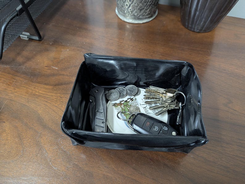 May include: A black, glossy, rectangular catch-all tray holding keys, coins, a folding knife, and a small decorative plate. The tray sits on a wooden surface, providing a convenient storage solution for everyday items.