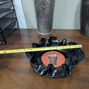 May include: A black, ruffled bowl made from a repurposed vinyl record. The bowl is approximately 10 inches in diameter and features a red and orange record label in the center. The bowl is on a wooden surface.