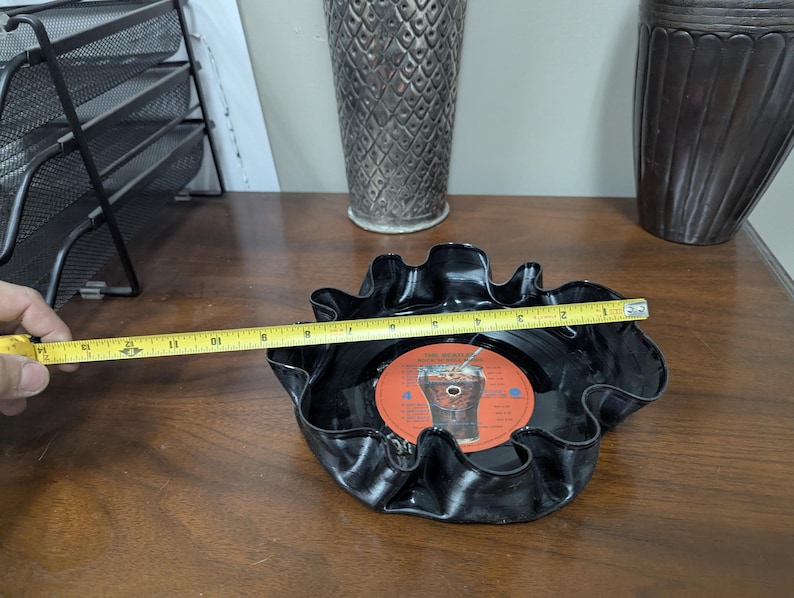 May include: A black, ruffled bowl made from a repurposed vinyl record. The bowl is approximately 10 inches in diameter and features a red and orange record label in the center. The bowl is on a wooden surface.