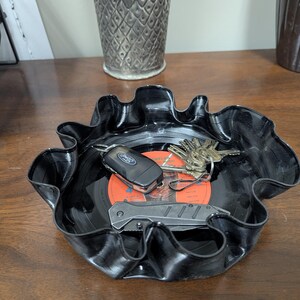 May include: A black, upcycled vinyl record bowl with a wavy edge. Inside the bowl are keys, a car key, and a small folding knife. The center of the record is orange with black text.