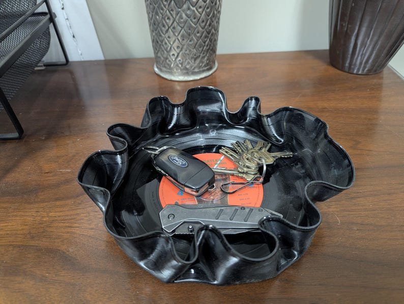 May include: A black, upcycled vinyl record bowl with a wavy edge. Inside the bowl are keys, a car key, and a small folding knife. The center of the record is orange with black text.