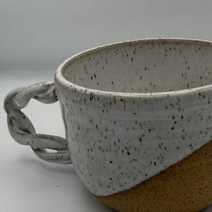 May include: A handmade ceramic mug with a twisted handle. The mug features a speckled white upper half and a tan lower half. The handle is a twisted rope design. The mug is designed for hot beverages.