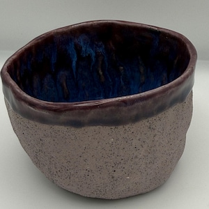Puede incluir: Un cuenco de cerámica hecho a mano con un exterior morado moteado y un interior azul profundo. El borde es de un tono morado más oscuro. El cuenco tiene una forma orgánica única, ideal para servir o exhibir.