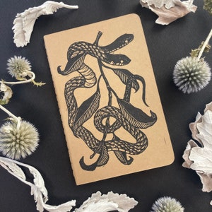 May include: A brown notebook featuring a black and white illustration of snakes entwined around a branch. The illustration is in a vintage style.