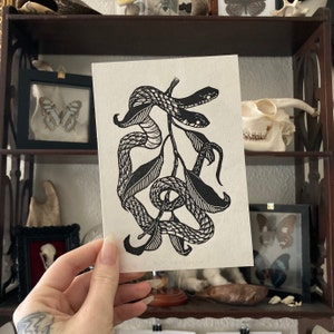 May include: Black and white linocut print of three snakes wrapped around a branch with leaves. The snakes are stylized and have a detailed pattern.