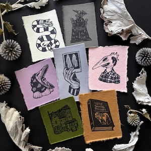 May include: A set of eight black and white linocut prints on different colored paper. The prints feature various spooky and gothic imagery, including a snake, a house, a hand holding a photo of ghosts, a creature with a beak, and a book titled "Handbook for the Recently Deceased".