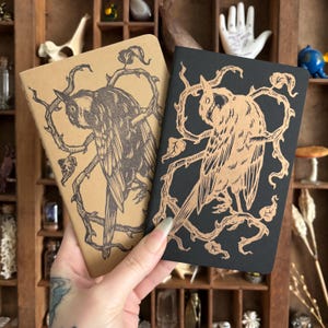 Peut inclure: Deux carnets, un beige et un noir, sont tenus dans une main. Chaque carnet présente une illustration détaillée d'un oiseau perché sur une branche avec des feuilles. Le motif de l'oiseau et de la branche est de couleur contrastante sur chaque carnet.
