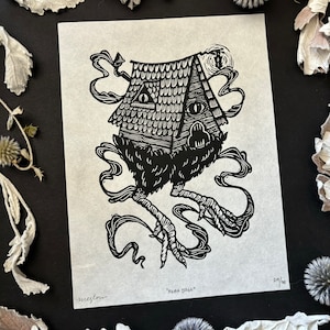May include: Black and white linocut print of a house with chicken legs, eyes, and smoke-like swirls. The print is titled "BABA YAGA" and is numbered 29/30. The artist's signature is in the lower left corner.