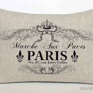 K&ouml;nnte beinhalten: Ein beigefarbenes Leinenkissen mit schwarzem Text, der "Marche Aux Puces PARIS No. 07, rue Jules Vall&egrave;s" lautet, mit einer Krone und einem floralen Design.