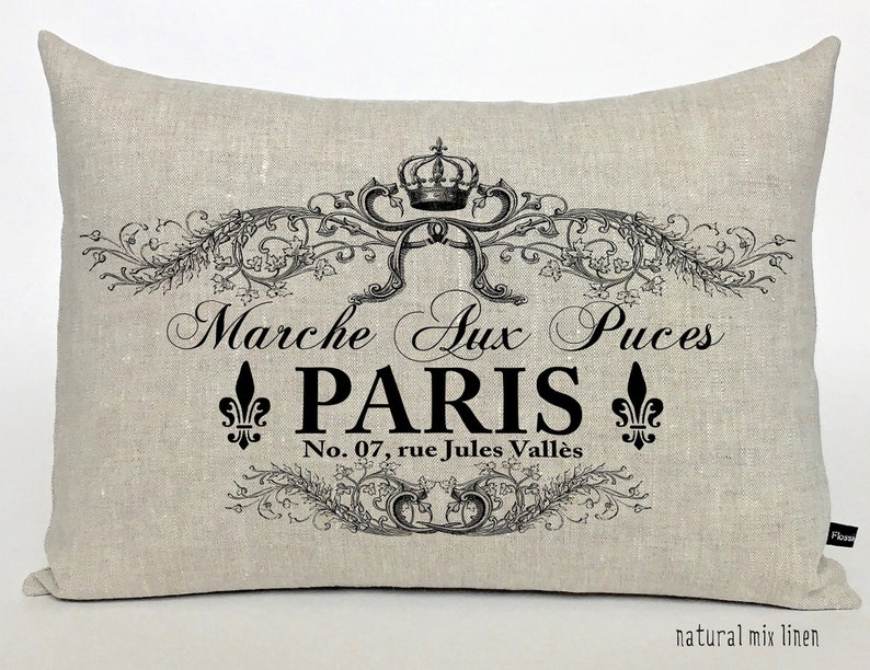 K&ouml;nnte beinhalten: Ein beigefarbenes Leinenkissen mit schwarzem Text, der "Marche Aux Puces PARIS No. 07, rue Jules Vall&egrave;s" lautet, mit einer Krone und einem floralen Design.