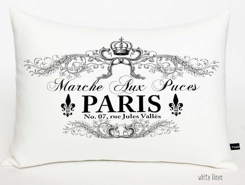 K&ouml;nnte beinhalten: Ein wei&szlig;es Leinenkissen mit einem schwarz-wei&szlig;en grafischen Design mit einer Krone, floralen Akzenten und dem Text "Marche Aux Puces PARIS No. 07, rue Jules Vall&egrave;s".