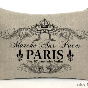 K&ouml;nnte beinhalten: Ein beigefarbenes Leinenkissen mit einem schwarz-wei&szlig;en Design mit einer Krone, floralen Akzenten und dem Text "Marche Aux Puces PARIS No. 07, rue Jules Vall&eacute;es".