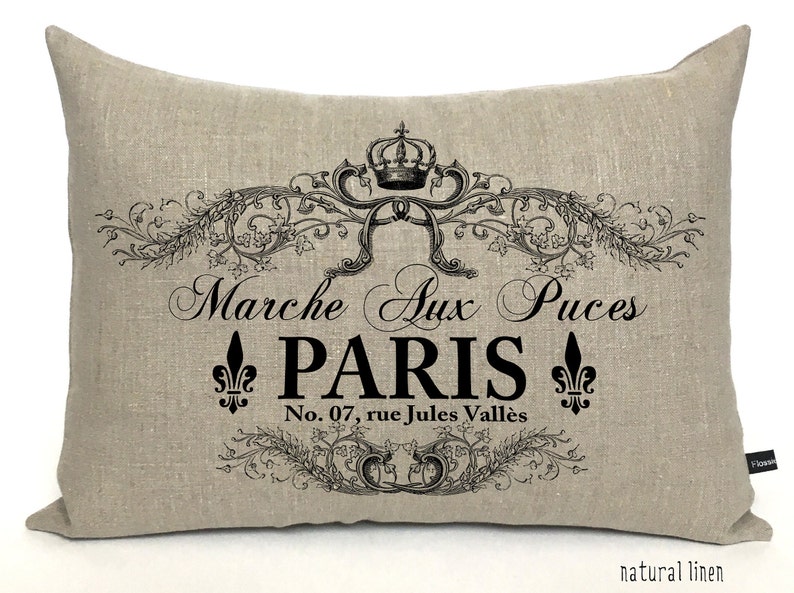 K&ouml;nnte beinhalten: Ein beigefarbenes Leinenkissen mit einem schwarz-wei&szlig;en Design mit einer Krone, floralen Akzenten und dem Text "Marche Aux Puces PARIS No. 07, rue Jules Vall&eacute;es".