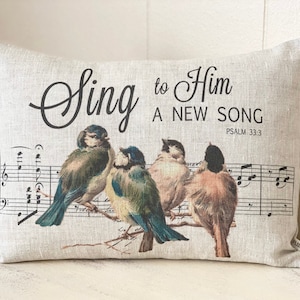 May include: A beige linen pillow with a black and white musical note design and four colorful birds. The text on the pillow reads "Sing to Him A New Song Psalm 33:3".