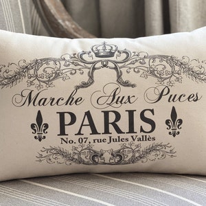 May include: A white pillow with black text and a decorative design. The text reads "Marche aux Puces PARIS No. 07, rue Jules Vallés".