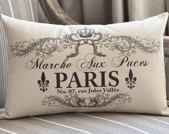 Puede incluir: Una almohada blanca con texto negro y un diseño decorativo. El texto dice "Marche aux Puces PARIS No. 07, rue Jules Vallés".