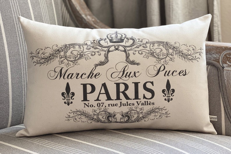 K&ouml;nnte beinhalten: Ein wei&szlig;es Kissen mit schwarzem Text und einem dekorativen Design. Der Text lautet "Marche aux Puces PARIS No. 07, rue Jules Vall&eacute;s".