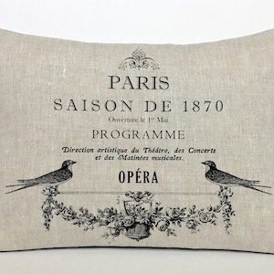 May include: A beige linen pillow with black text that reads "PARIS SAISON DE 1870 Ouverture le 1" Mai PROGRAMME Direction artistique du Théâtre, des Concerts et des Matinées musicales. OPÉRA" and a black and white illustration of two birds and floral designs.