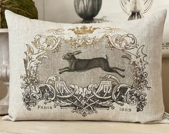 May include: A decorative pillow with a linen fabric cover features a vintage illustration of a running hare in a gold and black ornate frame. The text "Paris 1889" is printed at the bottom of the frame.