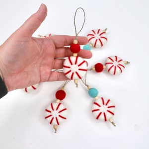 May include: A garland of felt peppermint ornaments with red and white stripes, strung with red and blue beads. The garland is hanging from a hand.