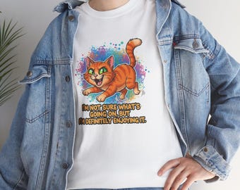 Puede incluir: Camiseta blanca con un gato atigrado naranja de dibujos animados con ojos verdes. El gato corre y sonríe, sobre un fondo de salpicaduras de colores. El texto de la camiseta dice: "I'M NOT SURE WHAT'S GOING ON, BUT I'M DEFINITELY ENJOYING IT."