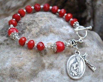 May include: Red beaded bracelet with silver-toned accents. The bracelet features a toggle clasp and a charm with a religious figure and a cross. The beads are faceted and round, with a bright red color.
