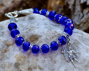 May include: A blue beaded bracelet with a silver clasp and a dangling angel charm. The faceted beads are a deep sapphire color, strung together with silver accents. The angel charm is detailed with outstretched wings.