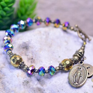May include: A gold and iridescent blue rosary bracelet with a Miraculous Medal charm. The bracelet features faceted glass beads and a toggle clasp.