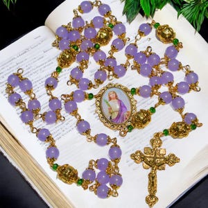 May include: A rosary featuring lavender-colored beads, gold-toned metal accents, and a central oval pendant with a religious image. The rosary includes a crucifix and is displayed on an open book.