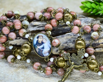 May include: A pink and white stone rosary with a bronze metal crucifix and a round pendant featuring a picture of Saint Anthony of Padua holding a baby.