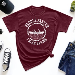 May include: A maroon t-shirt with a white graphic of a canoe with two people paddling and the text "Paddle Faster I Hear Banjos".